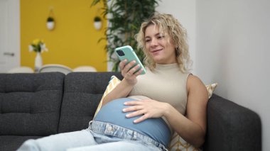 Genç hamile kadın akıllı telefon kullanıyor. Evde karnına dokunuyor.