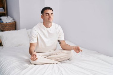 Young hispanic man doing yoga exercise sitting on bed at bedroom