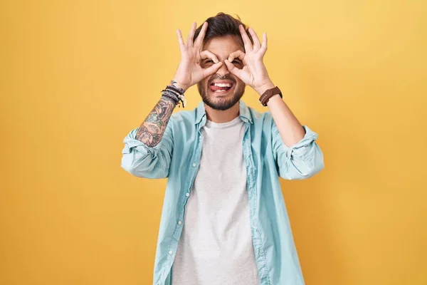 Young Hispanic Man Tattoos Standing Yellow Background Doing Gesture Binoculars — Fotografia de Stock