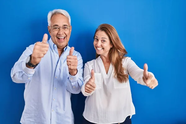 青い背景の上に立っている中年のパニックカップルは 手で積極的なジェスチャーを行うことを承認し 親指を上げて笑顔と成功のために幸せ 勝者のジェスチャー — ストック写真
