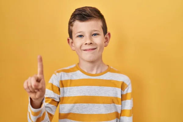 自信と幸せ笑みを浮かべている間 黄色の背景に立っている若い白人の子供は指の数1を示すと指摘します — ストック写真