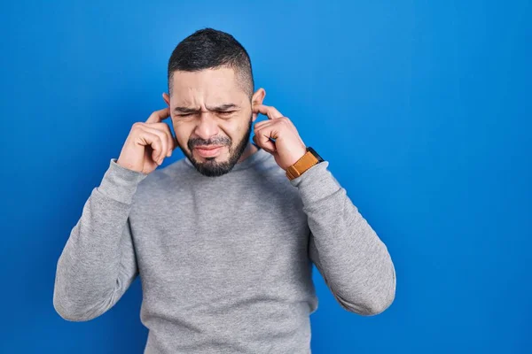 青い背景の上に立つヒスパニック系の男は 大きな音楽のノイズのためのいらいらした表情で指で耳を覆う 聴覚障害の概念 — ストック写真