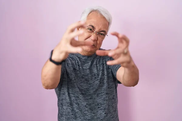 Middle age man with grey hair standing over pink background shouting frustrated with rage, hands trying to strangle, yelling mad