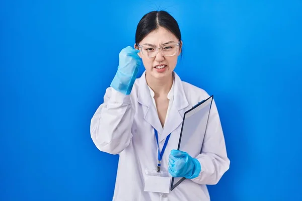 Chinese young woman working at scientist laboratory angry and mad raising fist frustrated and furious while shouting with anger. rage and aggressive concept.