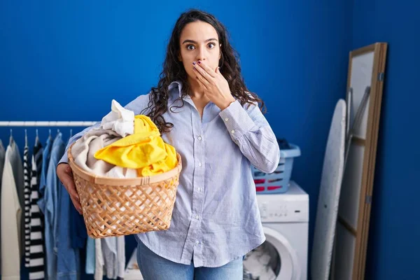 Young Brunette Woman Holding Laundry Basket Covering Mouth Hand Shocked — Stock Photo, Image