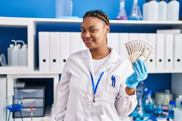 在科学家实验室工作的非洲裔美国妇女头戴辫子 手里拿着钱 站在那里 面带微笑 面带微笑 看上去很积极 很快乐 — 图库照片