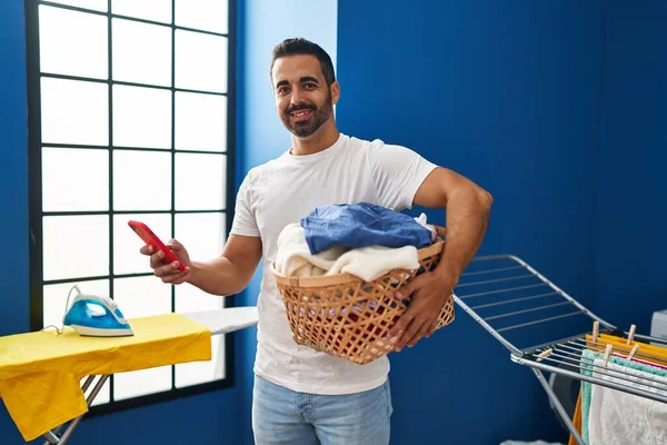 Young Hispanic Man Holding Basket Clothes Using Smartphone Laundry Room — Stockfoto