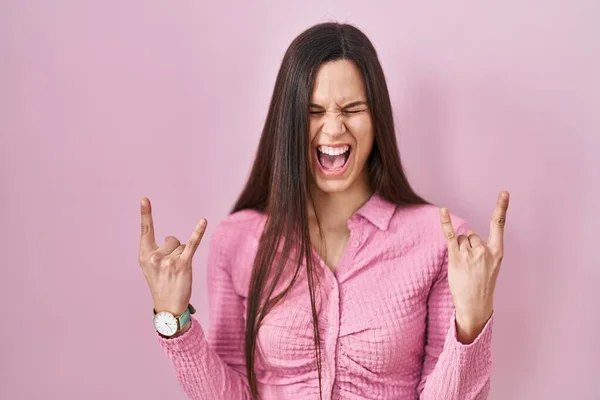 ピンクの背景の上に立つ若いヒスパニック系の女性が手を上げて岩のシンボルをやって狂気の表情で叫んでいます 音楽スター 重い音楽の概念は — ストック写真