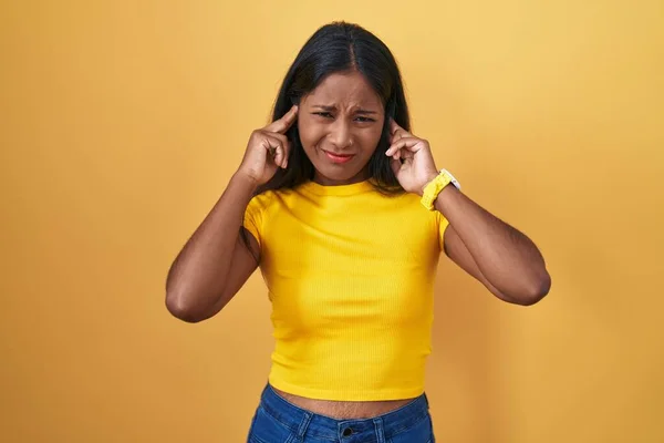 黄色い背景の上に立つ若いインド人女性は 大きな音楽のノイズに悩まされた表情で指で耳を覆います 聴覚障害の概念 — ストック写真