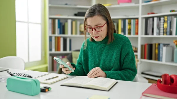 在图书馆用智能手机阅读一本书的成熟的灰发女学生 — 图库照片