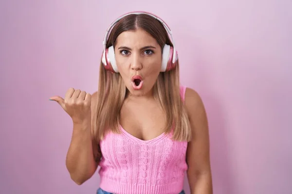 若いですブロンド女性を使用して音楽に耳を傾けますヘッドフォン驚くべきポインティングとともに手指で側面 オープン口驚きの表現 — ストック写真
