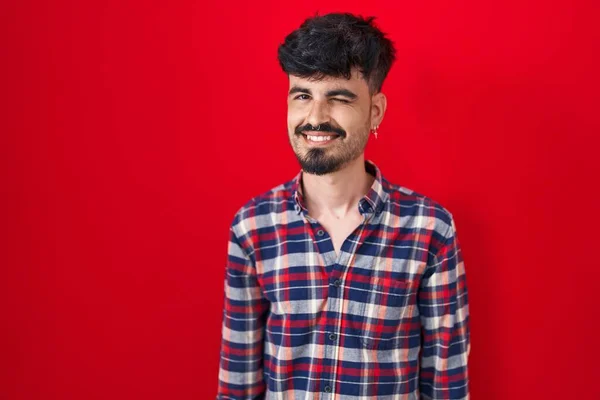 Joven Hombre Hispano Con Barba Pie Sobre Fondo Rojo Guiñando — Foto de Stock