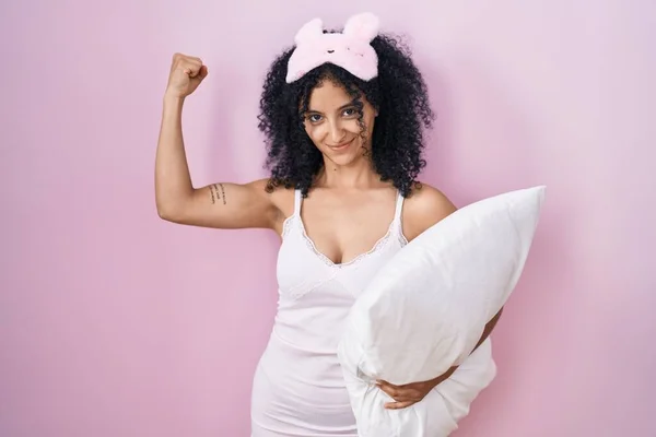 stock image Hispanic woman with curly hair wearing sleep mask and pajama holding pillow strong person showing arm muscle, confident and proud of power 