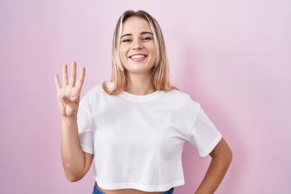 年轻的金发女人站在粉红的背景上 用四号手指指指着 自信而快乐地微笑着 — 图库照片