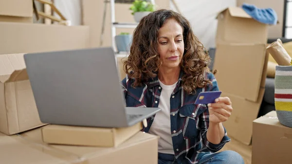 Middle age hispanic woman shopping with laptop and credit card sitting on floor at new home