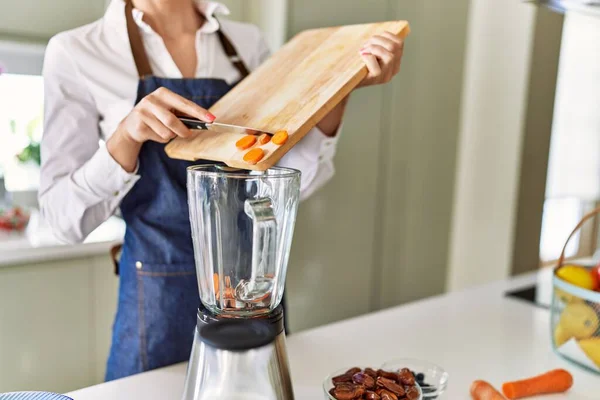 年轻的金发女人在厨房把胡萝卜倒在搅拌机上 — 图库照片