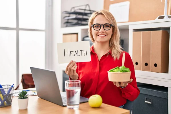 Blonde woman working at the office eating healthy food smiling with a happy and cool smile on face. showing teeth.