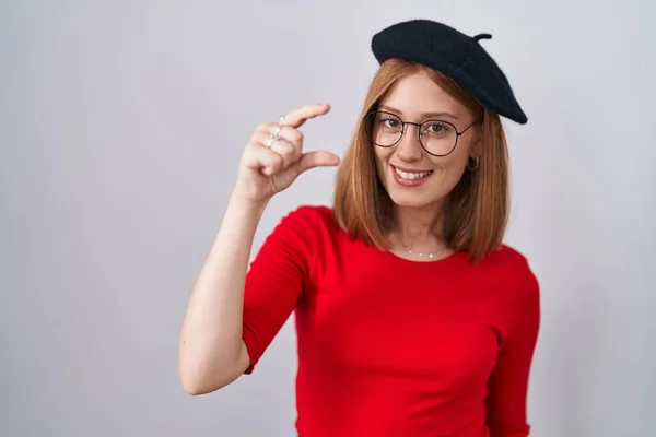 年轻的红头发女人站在那里 戴着眼镜 头戴贝雷帽 微笑着 自信地摆姿势 手拿着小尺寸的标志 手拿着手指 手拿着相机 计量概念 — 图库照片