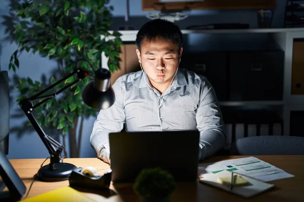 夜にコンピュータのノートパソコンを使って仕事をしている若い中国人男性が面白い顔で頬をむいている 口は空気で膨らみ狂気の表情で — ストック写真