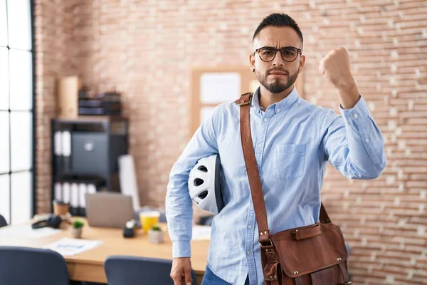 Young hispanic man working at the office holding bike helmet annoyed and frustrated shouting with anger, yelling crazy with anger and hand raised