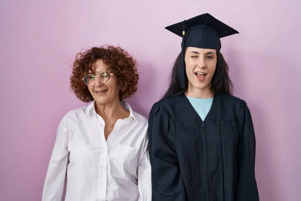 ヒスパニック系の母親と娘を身に着けています卒業キャップと式のローブウインク見ますザ カメラとともにセクシー表現 陽気で幸せな顔 — ストック写真