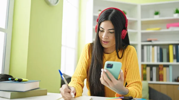 図書館大学でスマートフォンやヘッドフォンを使ってノートを書く若い美しいヒスパニック系女性の学生 — ストック写真