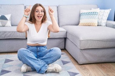 Genç beyaz kadın oturma odasında yerde oturuyor. Şaşırmış, şaşırmış ve parmaklarıyla işaret ediyor ve kollarını kaldırıyor.. 