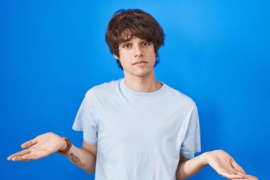 Hispanic young man standing over blue background clueless and confused with open arms, no idea concept. 