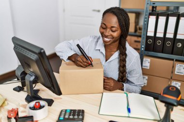 Afrika kökenli Amerikalı kadın ecommerce işçisi ofiste paket üzerinde bilgisayar kullanıyor.