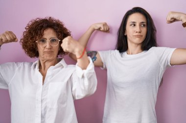 İspanyol anne ve kızı pembe arka planda gelişigüzel beyaz tişört giyiyorlar. Kollarında gururla gülümseyen kaslar var. fitness konsepti. 