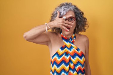 Sarı arka planda gri saçlı orta yaşlı bir kadın. Şoka girmiş, yüzünü ve gözlerini kapamış, utanmış bir ifadeyle parmaklarının arasından bakıyor.. 