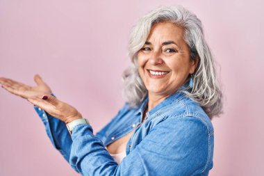 Pembe arka planda duran gri saçlı orta yaşlı bir kadın avuç içi açık bir şekilde fotokopi alanını gösteriyor, gülümseyerek mutlu bir gülümseme sergiliyor. 