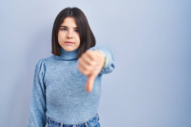 Mavi arka planda duran Latin kökenli genç kadın mutsuz ve kızgın görünüyor. Reddedilmiş ve olumsuz bir hareket sergiliyor. kötü ifade. 