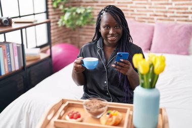 Afro-Amerikalı bir kadın yatak odasında akıllı telefon kullanarak kahvaltı yapıyor.