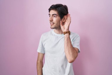 Young hispanic man standing over pink background smiling with hand over ear listening an hearing to rumor or gossip. deafness concept. 