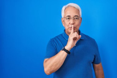 Middle age man with grey hair standing over blue background asking to be quiet with finger on lips. silence and secret concept. 