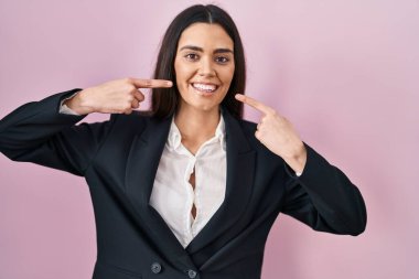 Genç esmer kadın pembe arka planda iş stili giyinmiş neşeli gülümseyen ve parmaklarıyla ve ağzıyla işaret eden. diş sağlığı kavramı. 