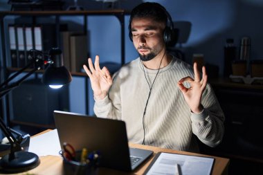 Genç yakışıklı adam geceleri bilgisayar kullanarak çalışıyor rahatla ve gözleri kapalı gülümse parmaklarıyla meditasyon hareketi yapıyor. yoga konsepti. 