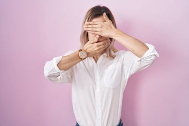 Pembe arka planda duran genç ve güzel bir kadın gözlerini ve ağzını ellerle kapatıyor, şaşırmış ve şok olmuş. duygu gizleme 