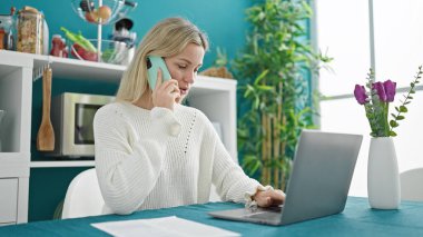 Genç sarışın kadın yemek odasında dizüstü bilgisayar kullanıyor.