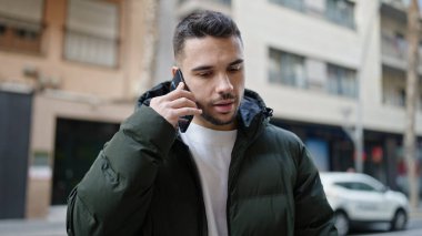 Young hispanic man talking on smartphone with serious expression at street