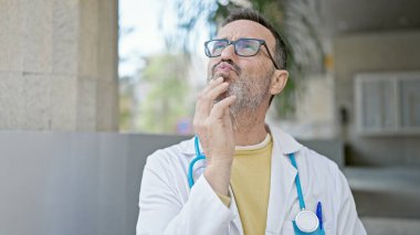 Orta yaşlı doktor şüpheli bir ifadeyle ayakta duruyor ve hastanede düşünüyor.