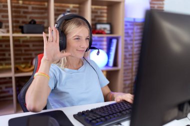 Video oyunları oynayan, kulaklık takan, mutlu, güler yüzlü, dostça bir jest yapan genç beyaz bir kadın. 