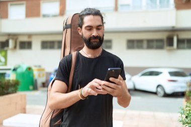 Young hispanic man musician using smartphone holding guitar case at park