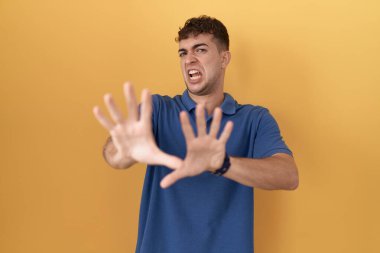 Sarı arka planda duran İspanyol genç adam korku ifadesinden korkmuş ve dehşete düşmüş. El hareketlerini durdur, şok içinde bağır. panik konsepti. 