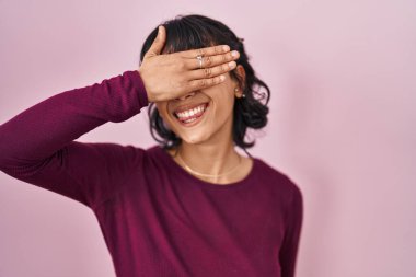 Pembe arka planda duran genç ve güzel bir kadın. Gülümsüyor ve gülümsüyor. Gözleri şaşkınlıkla örtüyor. kör kavram. 