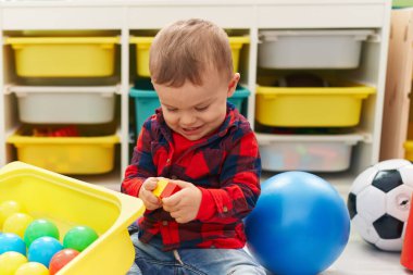 Anaokulunun zemininde oturan, taşaklarıyla oynayan sevimli sarışın çocuk.
