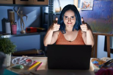 Mavi saçlı genç modern kız gece vakti dizüstü bilgisayarla resim stüdyosunda oturmuş mutlu ve komik yüzüyle kamerayı işaret ediyor. iyi enerji ve titreşimler. 