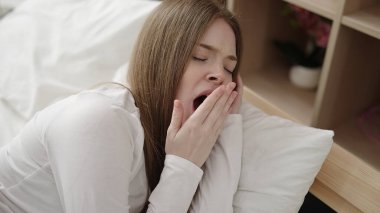 Young blonde woman lying on bed yawning at bedroom