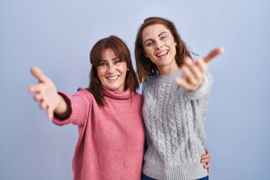Anne ve kızı mavi arka planda durup kucaklaşmak için kollarını açıp kameraya bakıyorlar. Mutluluğu kucaklayan neşeli bir ifade.. 